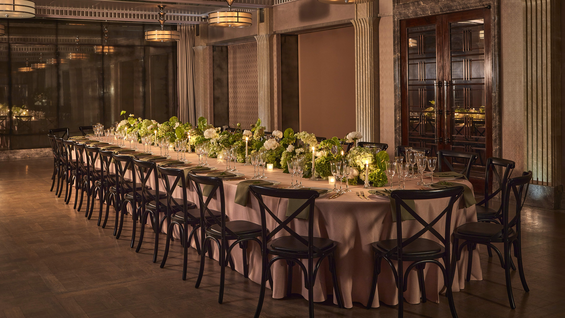 A sophisticated indoor dining setup featuring a long, elegant table adorned with lush green and white floral arrangements, glowing candles, and refined dark wood chairs in a softly lit room.