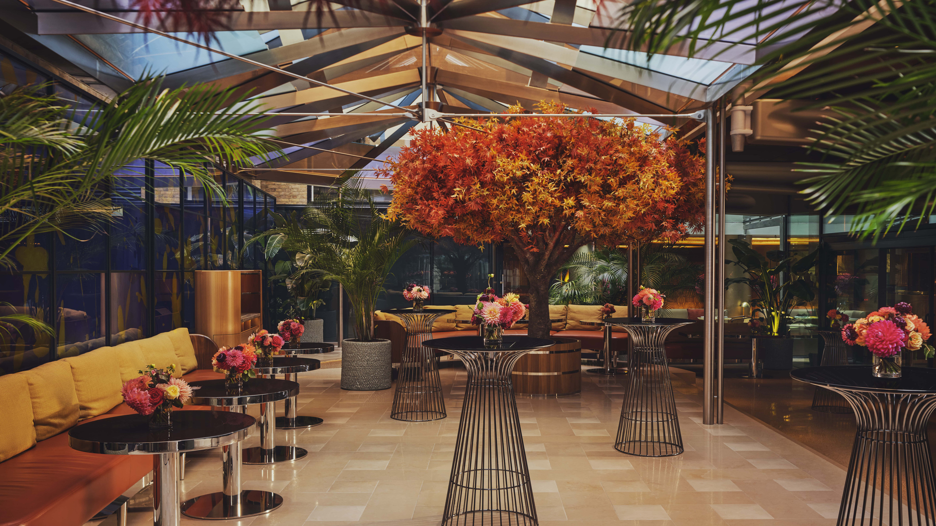 A contemporary glass-enclosed space featuring a vibrant orange maple tree centerpiece, surrounded by chic lounge furniture, sleek black tables, and bursts of colorful floral arrangements.