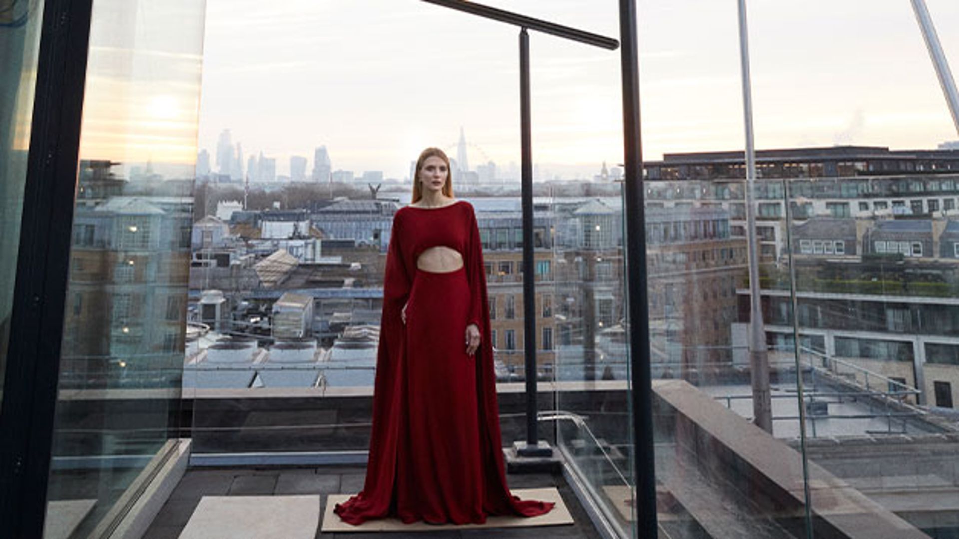 A woman in a striking red gown with a cut-out detail stands on The Emory's rooftop, overlooking London's skyline at sunset.