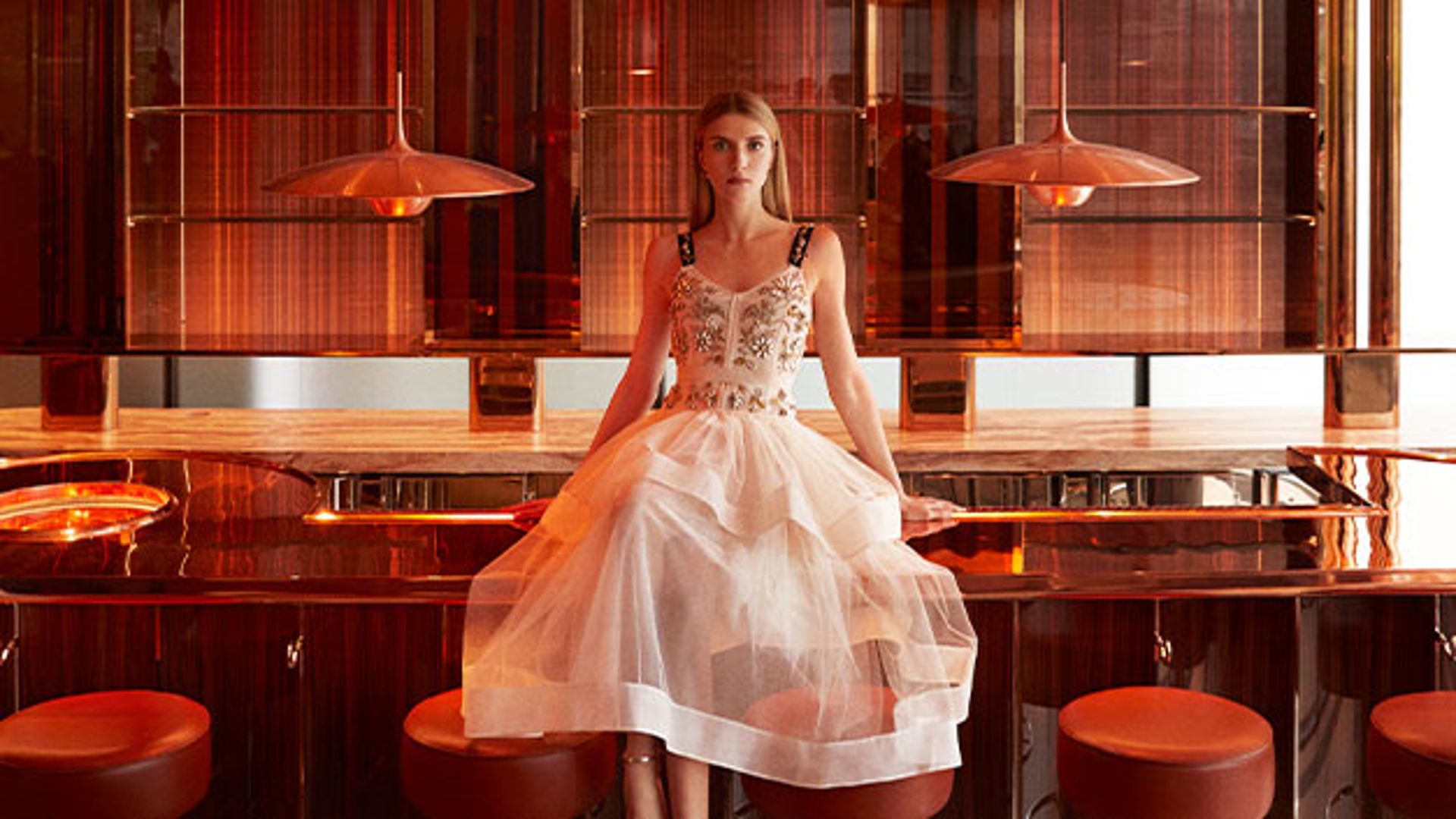 A woman in a delicate tulle dress sits on the counter of The Emory's amber-toned bar, surrounded by warm, modern decor.