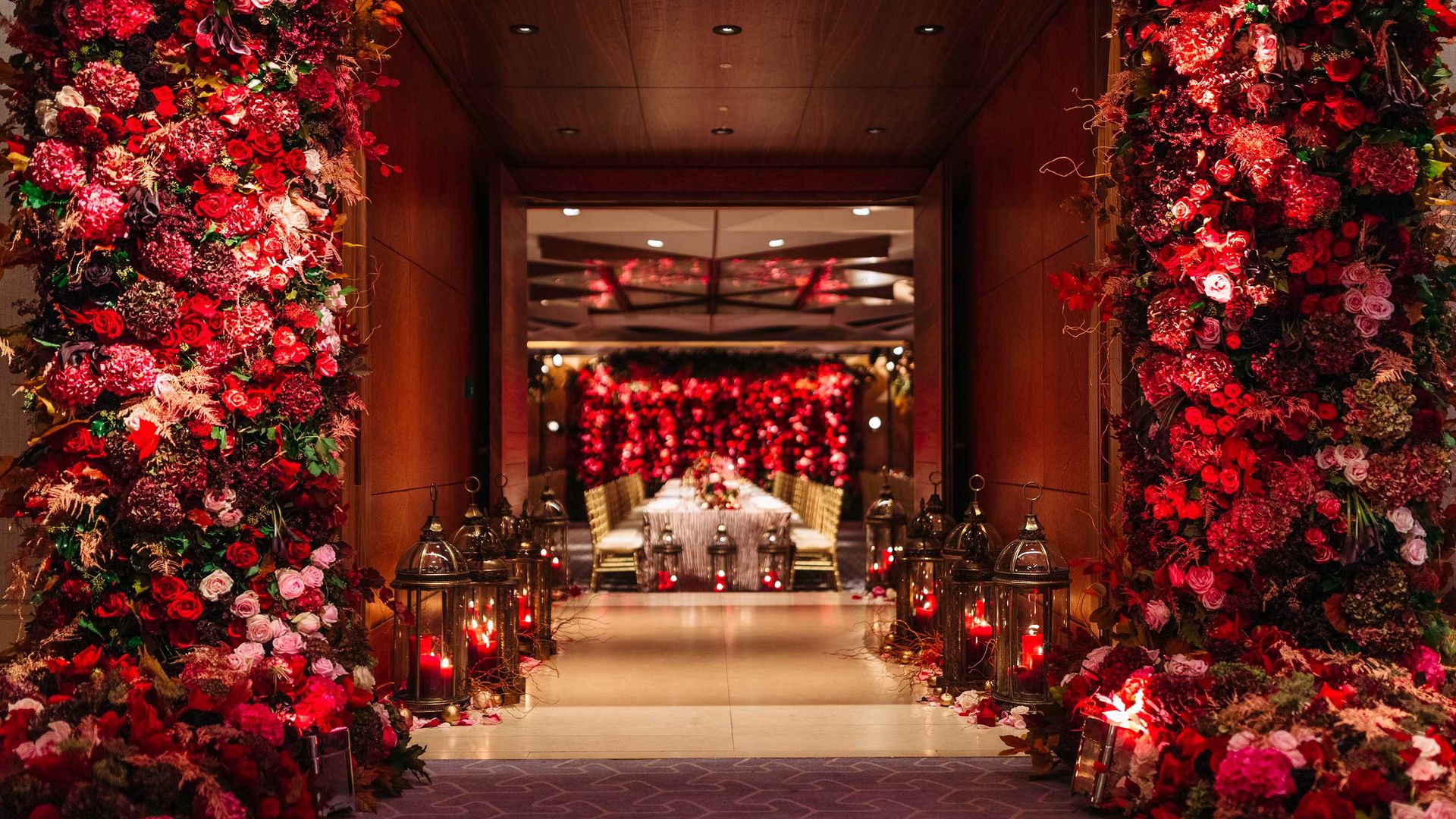 A dramatic entrance framed by cascading red and pink floral arrangements leads to a luxurious dining space illuminated by lanterns and soft, romantic lighting.