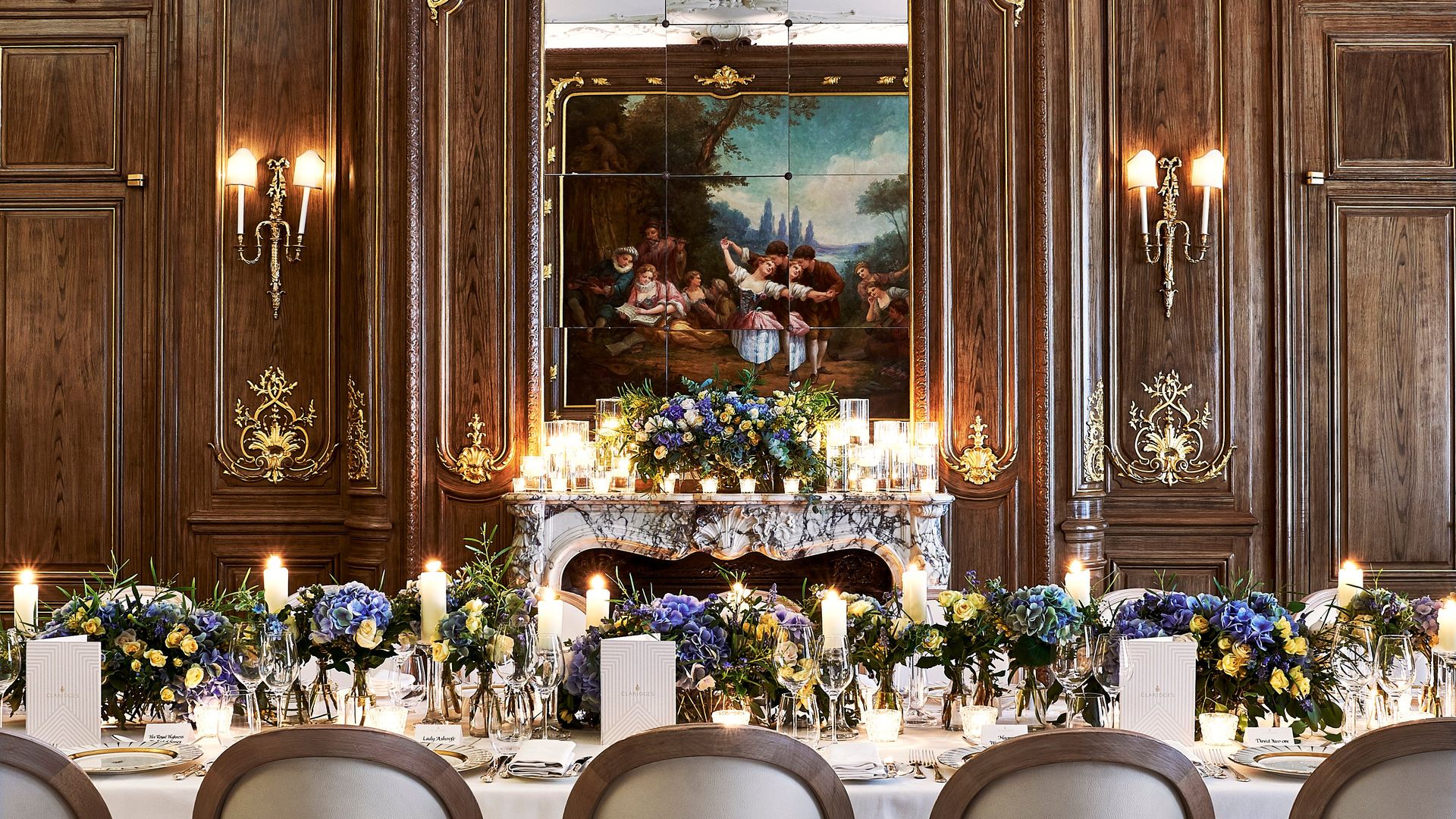 A luxurious banquet space showcasing an intricately detailed wood-paneled wall with gold embellishments, a grand marble fireplace, and a beautifully set table with vibrant floral arrangements and candlelight.