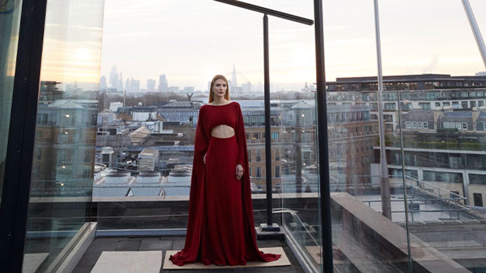 A woman in a striking red gown with a cut-out detail stands on The Emory's rooftop, overlooking London's skyline at sunset.