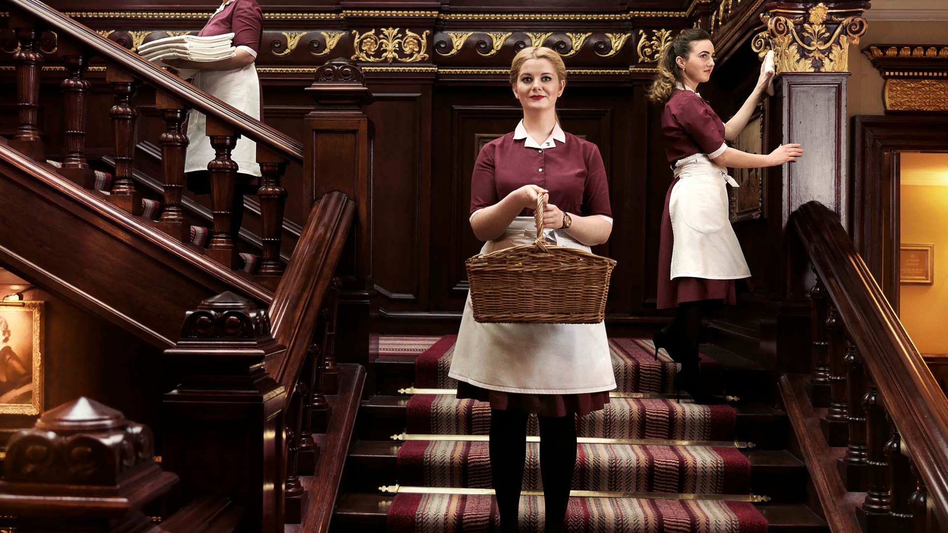 Three housekeepers in uniform work on a grand wooden staircase, carrying linens, dusting, and holding a wicker basket.