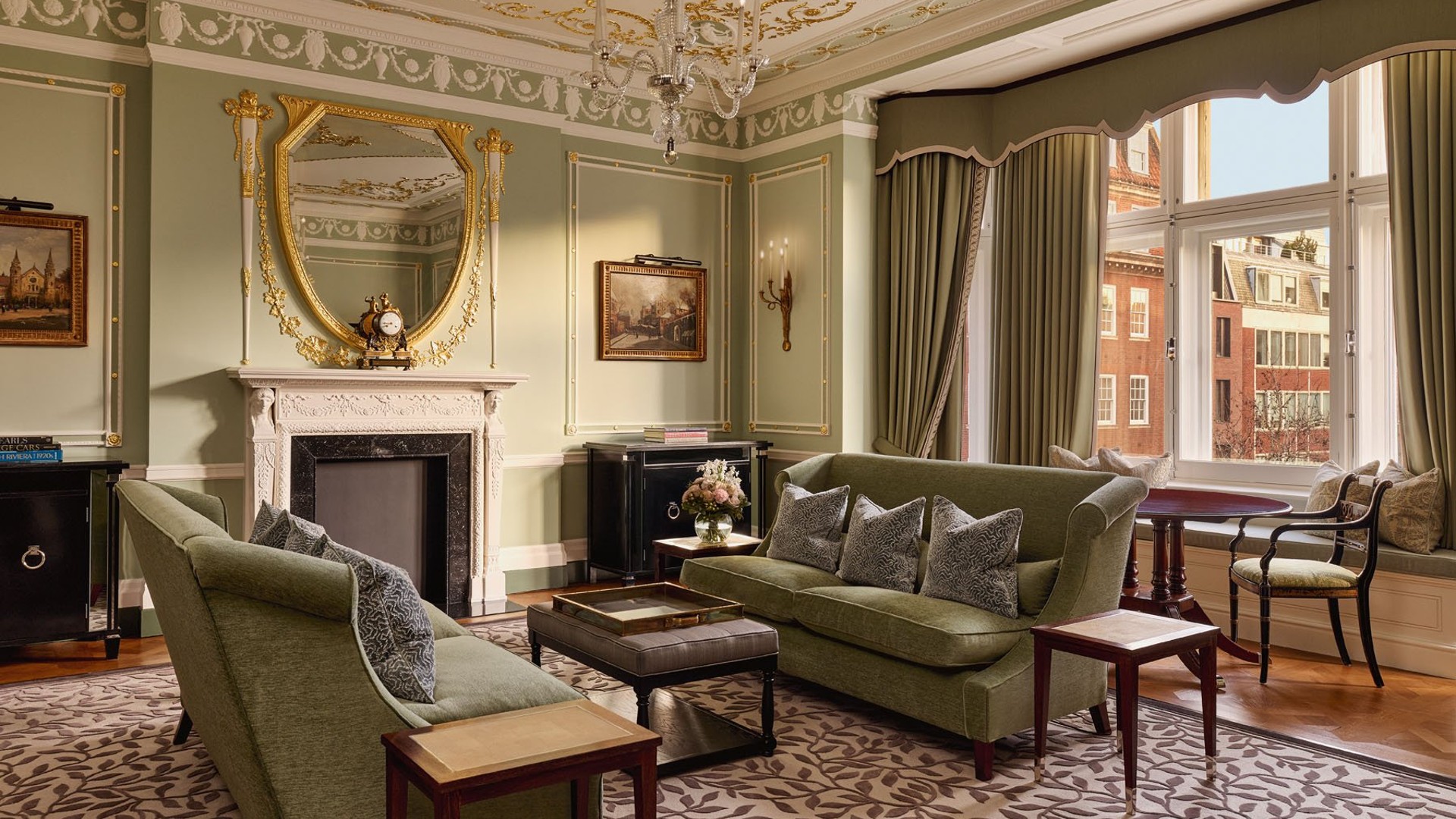 Elegant sitting room with green sofas, ornate ceiling, gilded mirror above fireplace, and large windows overlooking Mayfair buildings.