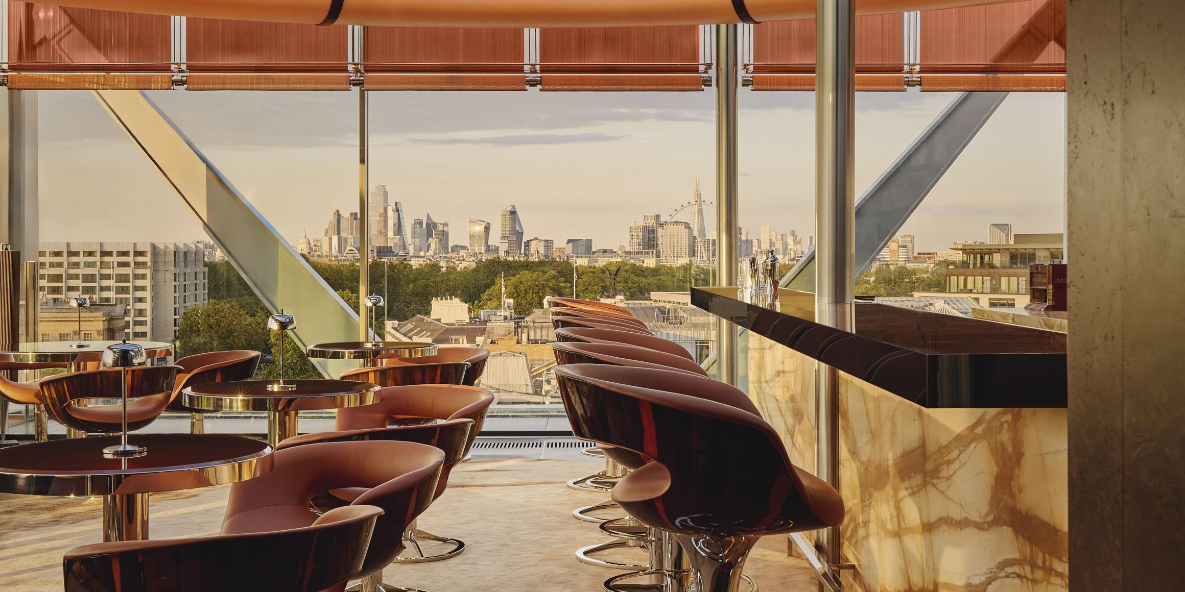 Stylish rooftop bar with leather stools, marble counter, and panoramic views of the London skyline through glass walls and ceiling.