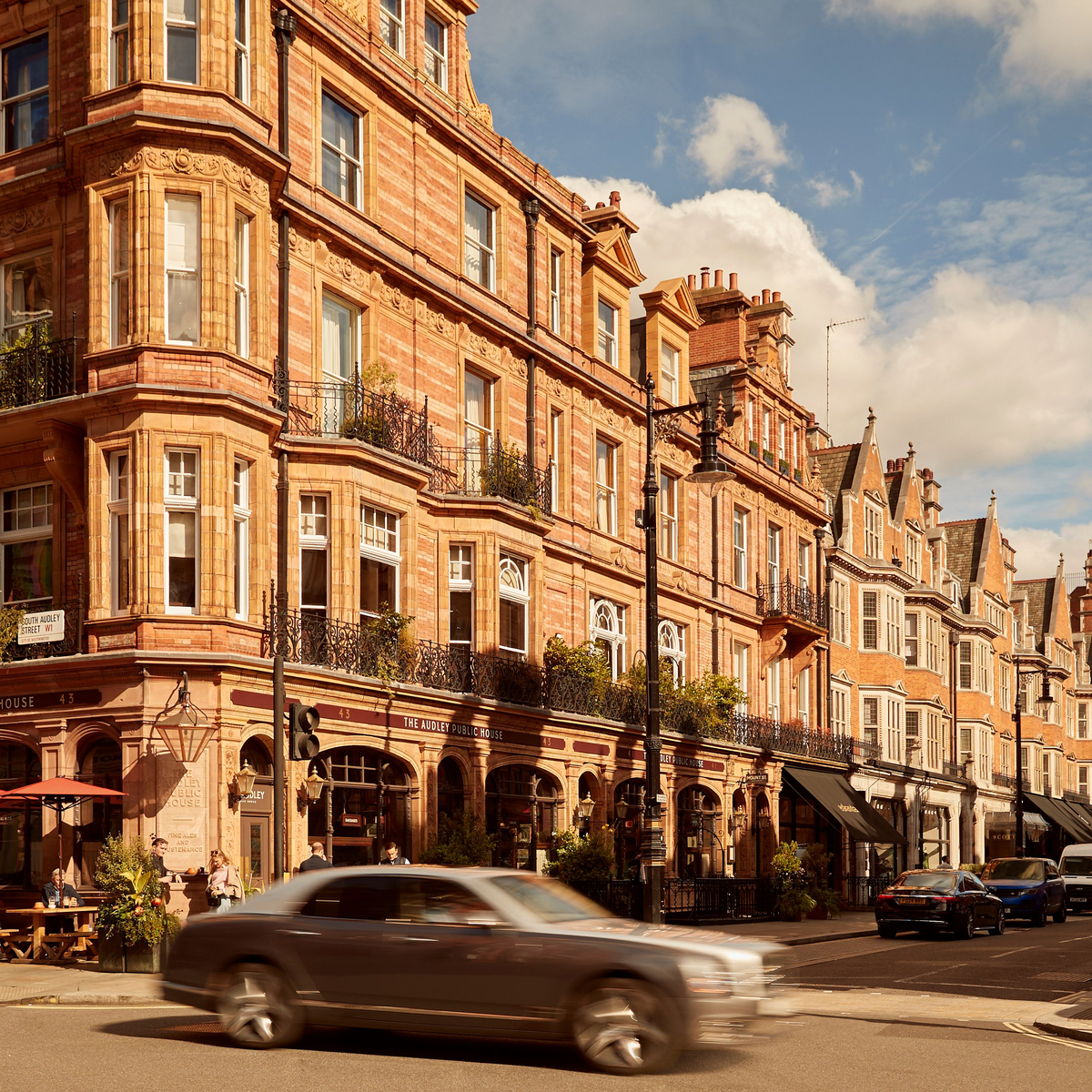 Charming street scene featuring historic red-brick architecture, bustling outdoor cafes, and passing cars under a sunny blue sky.