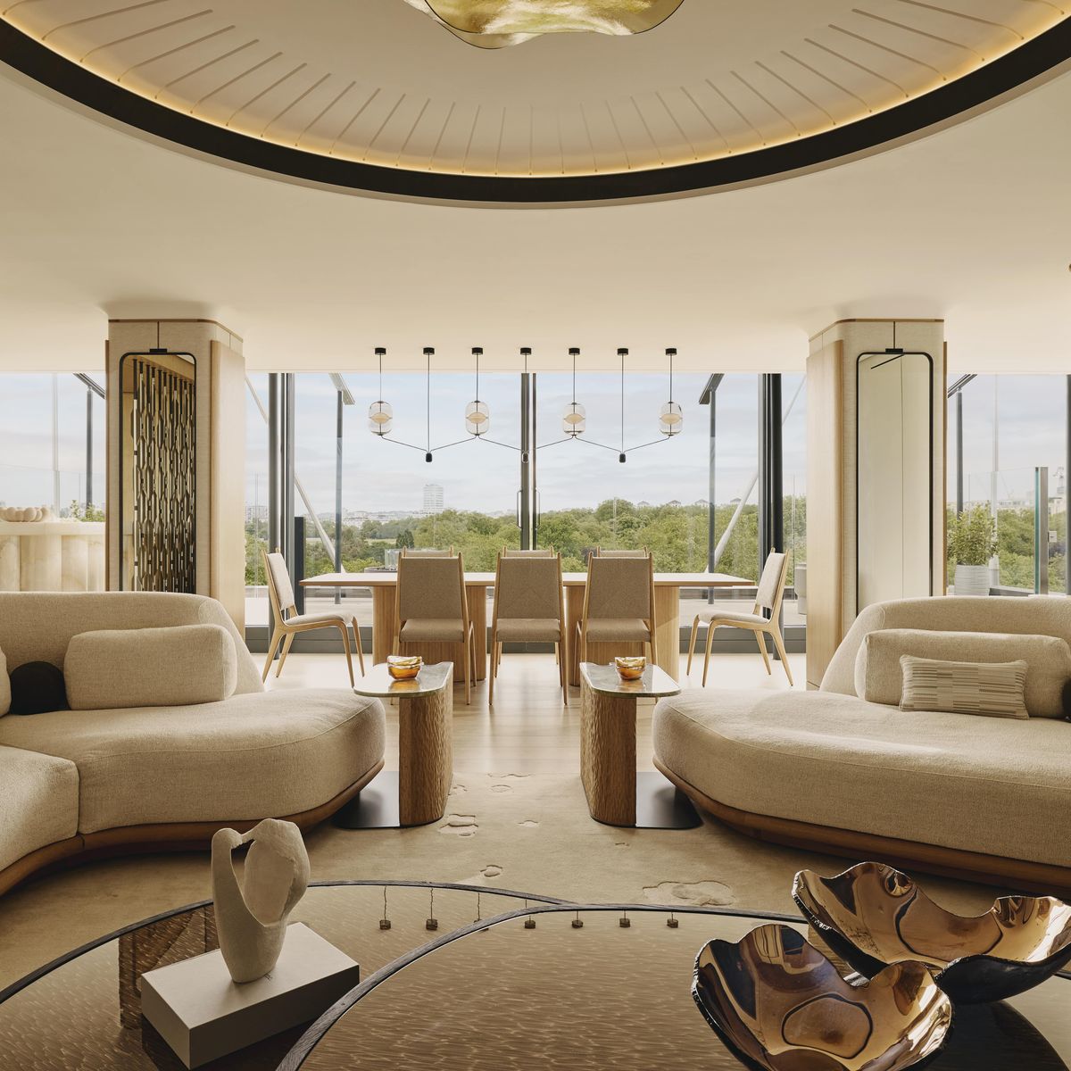 A modern, light-filled penthouse with curved sofas, a dining area, and floor-to-ceiling windows overlooking lush greenery.
