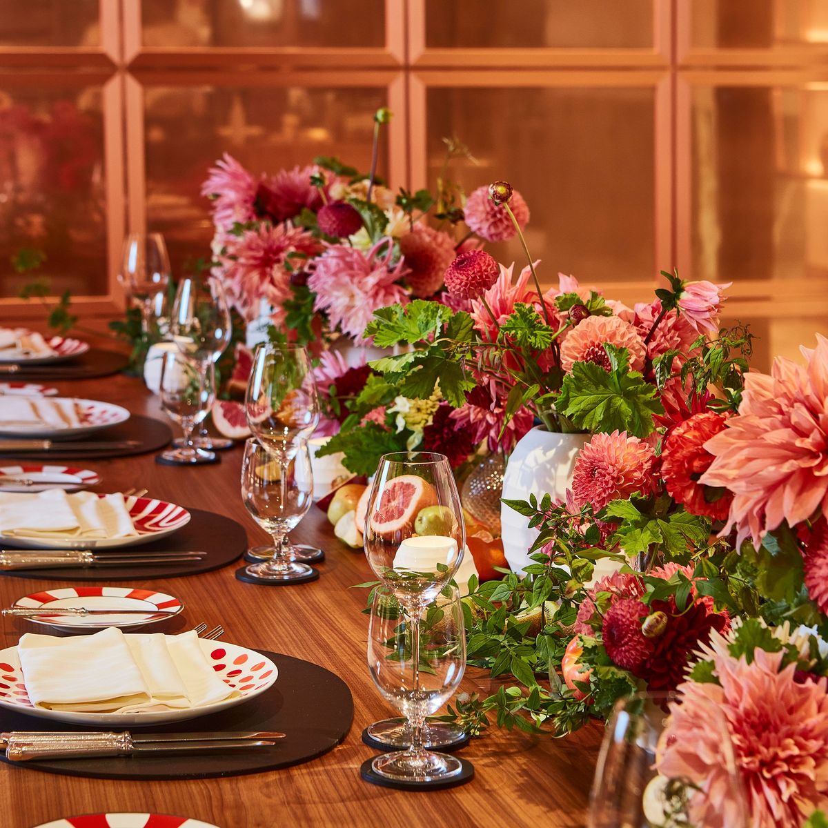 Close-up of a vibrant dining table set with red and white plates, crystal glasses, and lush pink floral arrangements with greenery and fresh fruit accents.