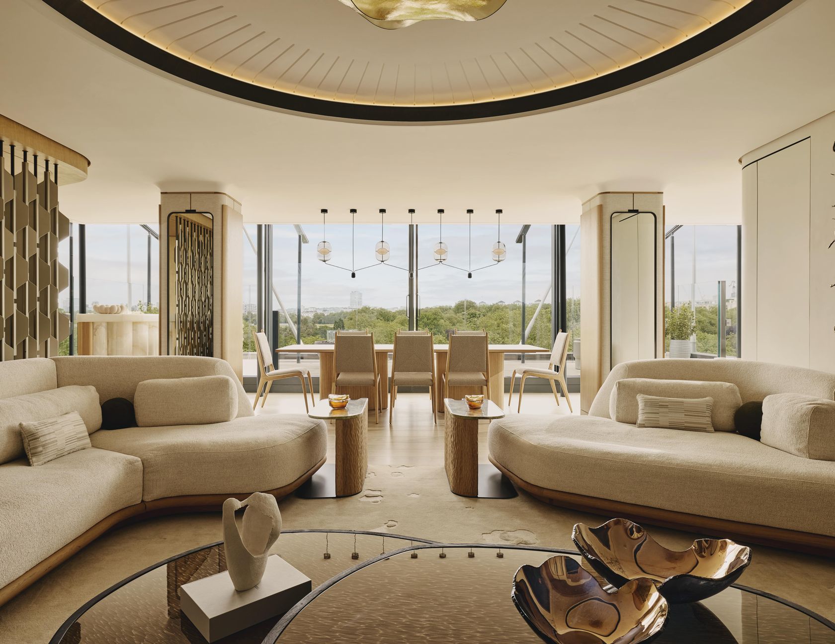 Curved cream sofas and sculptural décor in a modern penthouse living-dining space with floor-to-ceiling windows overlooking treetops and a distant skyline.