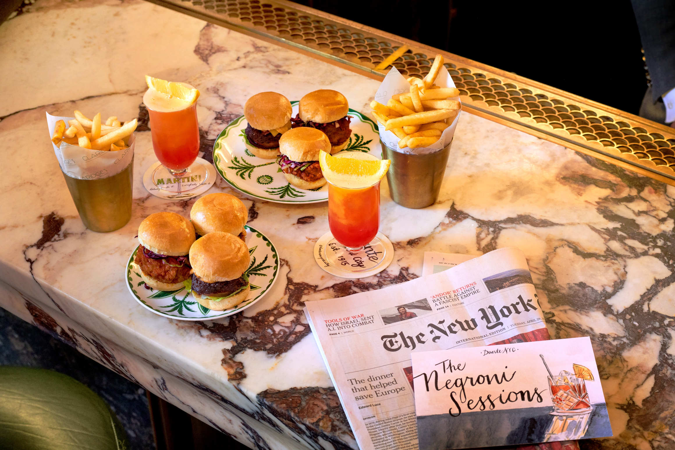 Sliders, fries, and cocktails sit on a marble bar beside a newspaper and “The Negroni Sessions” card.