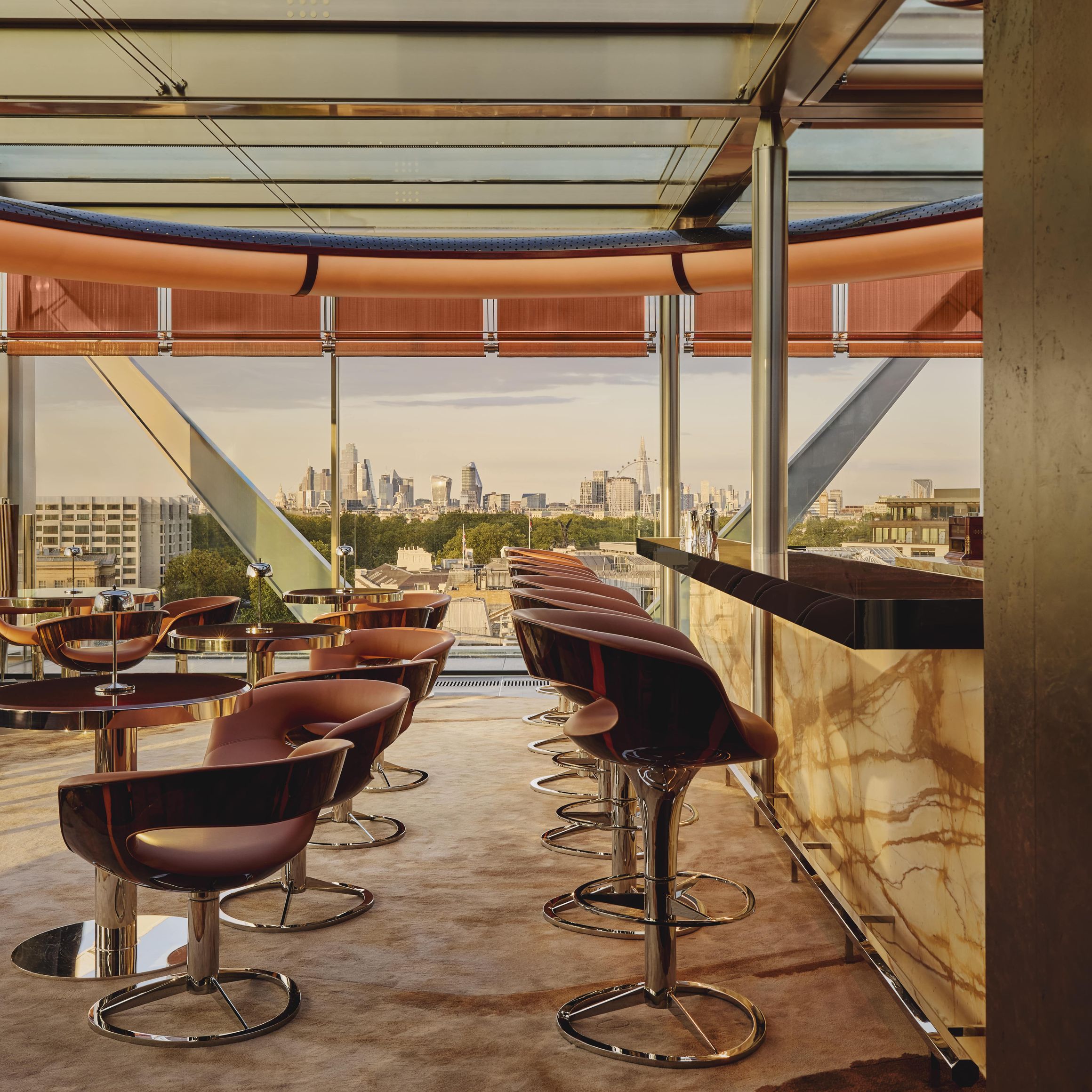 Stylish rooftop bar with leather stools, marble counter, and panoramic views of the London skyline through glass walls and ceiling.