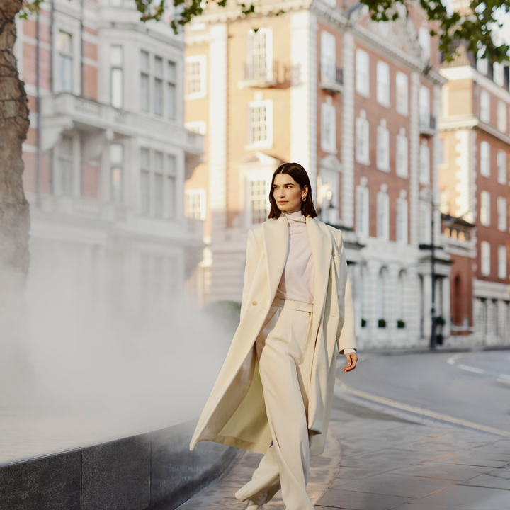 Elegant woman in a cream ensemble strolling through a refined urban setting, surrounded by classic architecture and soft mist.