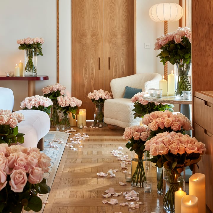 Luxurious room decorated with dozens of pink rose bouquets in glass vases and soft candlelight, with rose petals scattered along a parquet floor leading to a cozy white armchair beside a wooden door