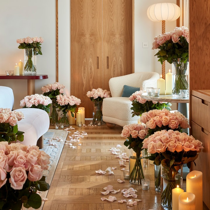 Luxurious room decorated with dozens of pink rose bouquets in glass vases and soft candlelight, with rose petals scattered along a parquet floor leading to a cozy white armchair beside a wooden door