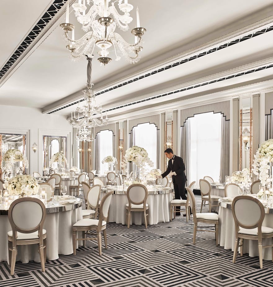 Elegant ballroom set for a wedding, with white floral centerpieces, chandeliers, and a waiter preparing tables.