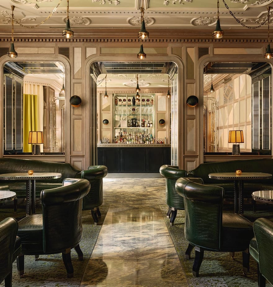 An elegant bar with dark green leather seating, marble floors, mirrored panels, and soft lighting, creating a refined, intimate ambiance.