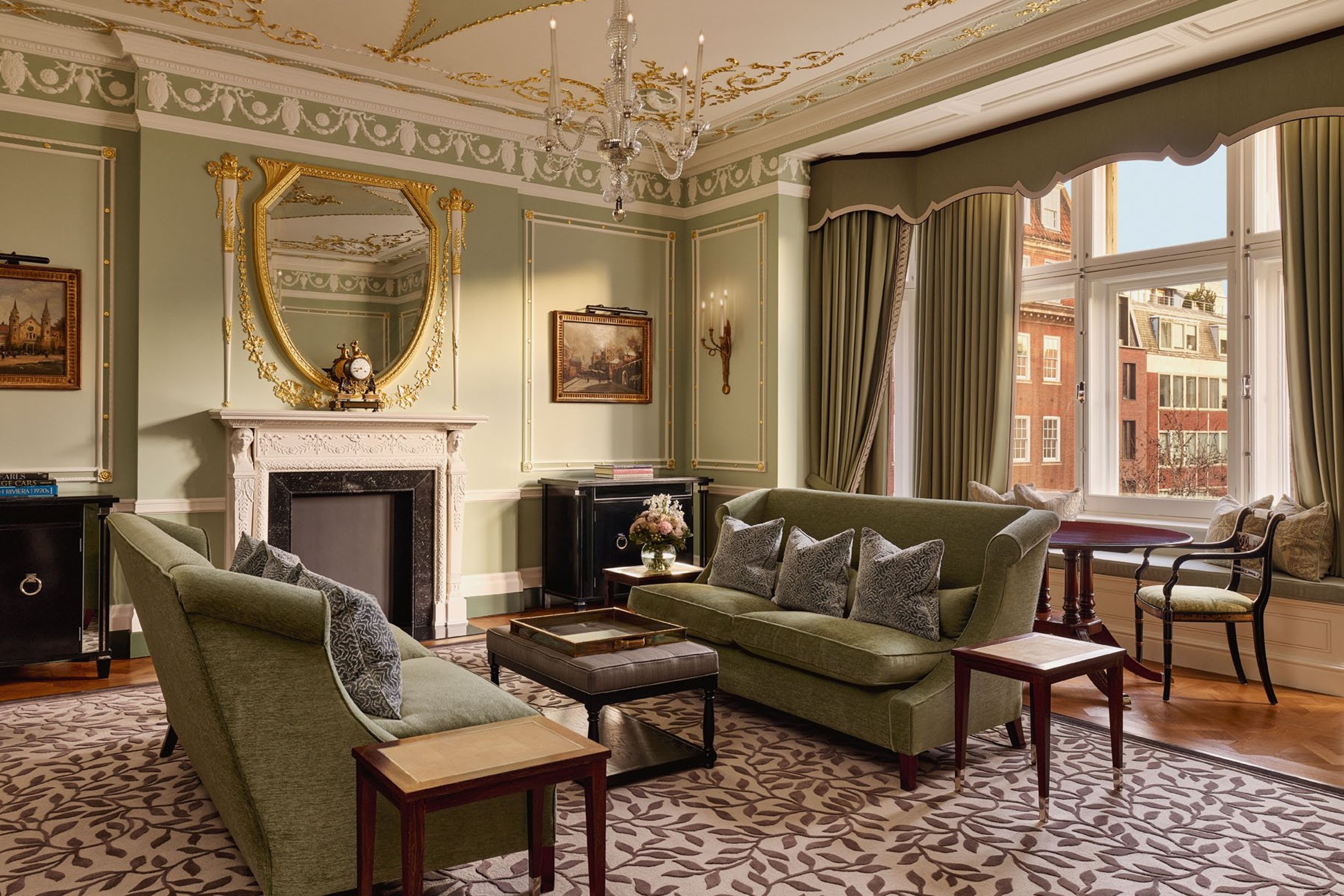 Elegant sitting room with green sofas, ornate ceiling, gilded mirror above fireplace, and large windows overlooking Mayfair buildings.