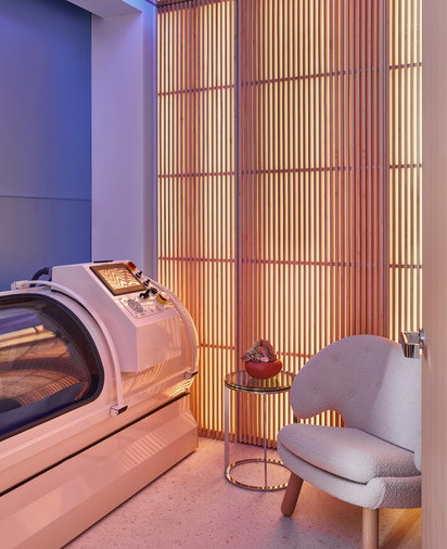 A sleek hyperbaric chamber in a softly lit wellness space, featuring warm wood accents, a plush chair, and calming ambiance.