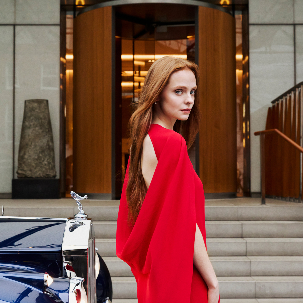 A woman in a flowing red dress stands beside a classic dark blue car outside a grand building entrance.