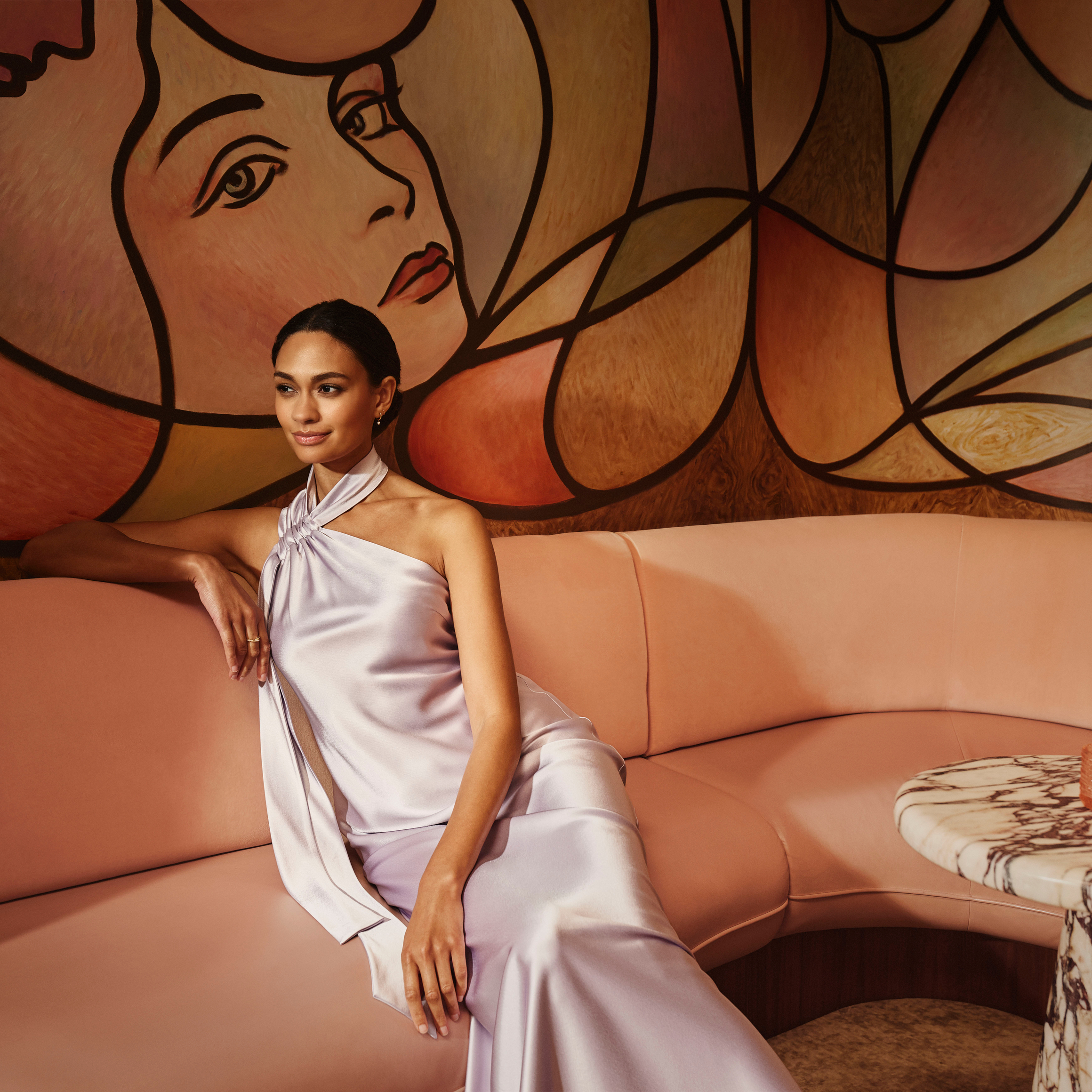 Elegant woman in a satin gown seated on a blush sofa, set against a mural with abstract, stylized faces.