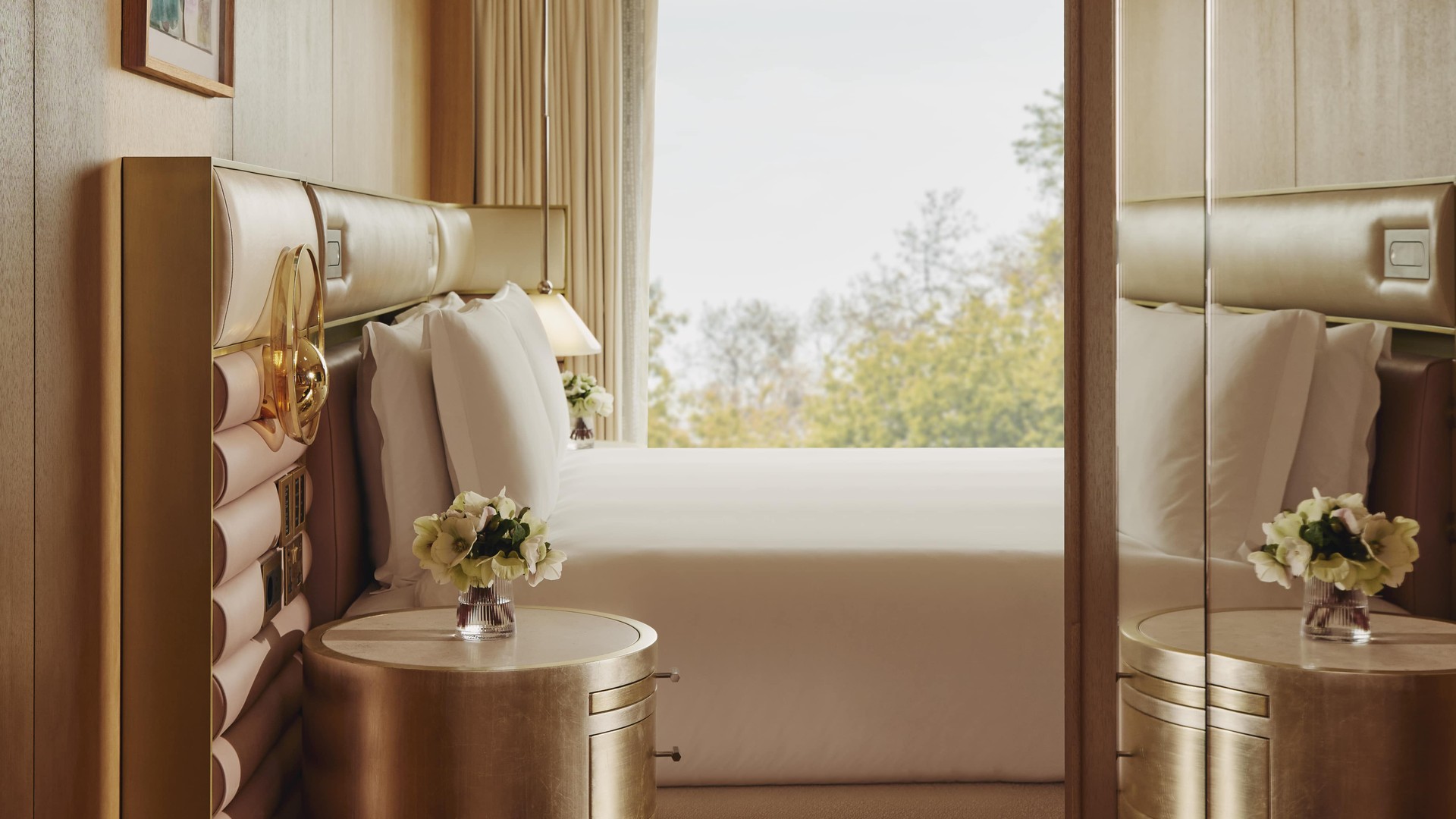 Luxurious bedroom with gold-trimmed headboard, mirrored panels, and large window overlooking treetops, evoking calm sophistication.