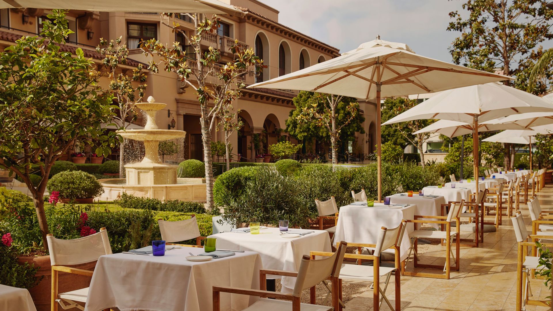 Elegant outdoor terrace dining with white tablecloths, large parasols, and a central stone fountain in manicured gardens.