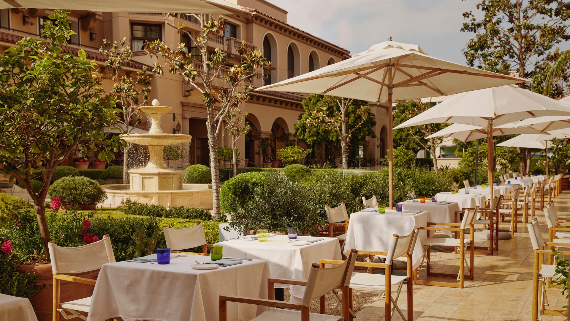 Elegant outdoor terrace dining with white tablecloths, large parasols, and a central stone fountain in manicured gardens.