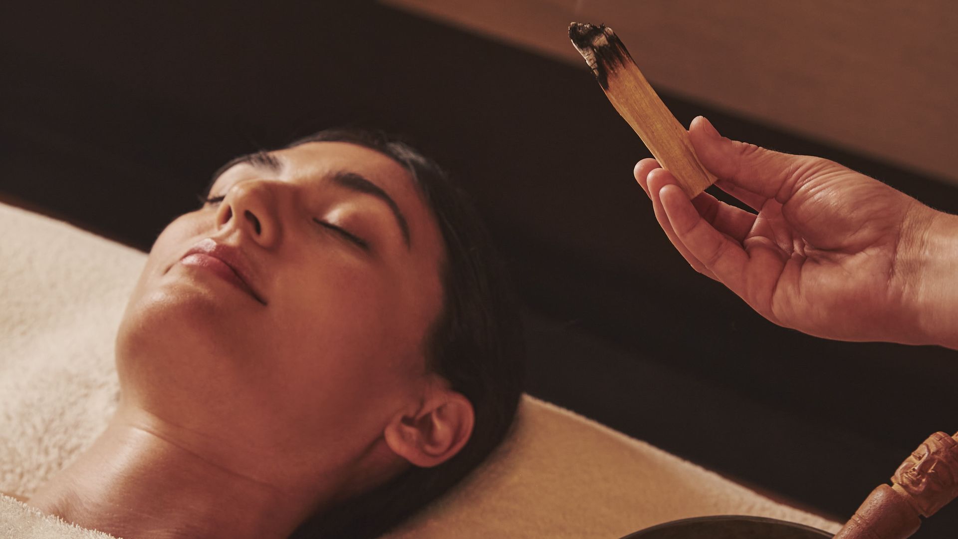 A peaceful spa scene with a person lying on a treatment table, eyes closed, as fragrant smoke rises from a ritual incense stick near a Tibetan singing bowl.