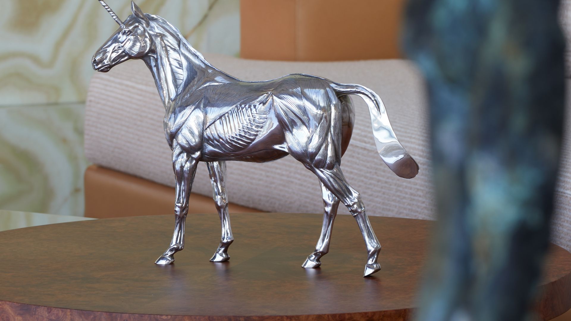 A sleek silver unicorn sculpture displayed on a rich wooden table against a backdrop of neutral-toned furniture and textured stone.