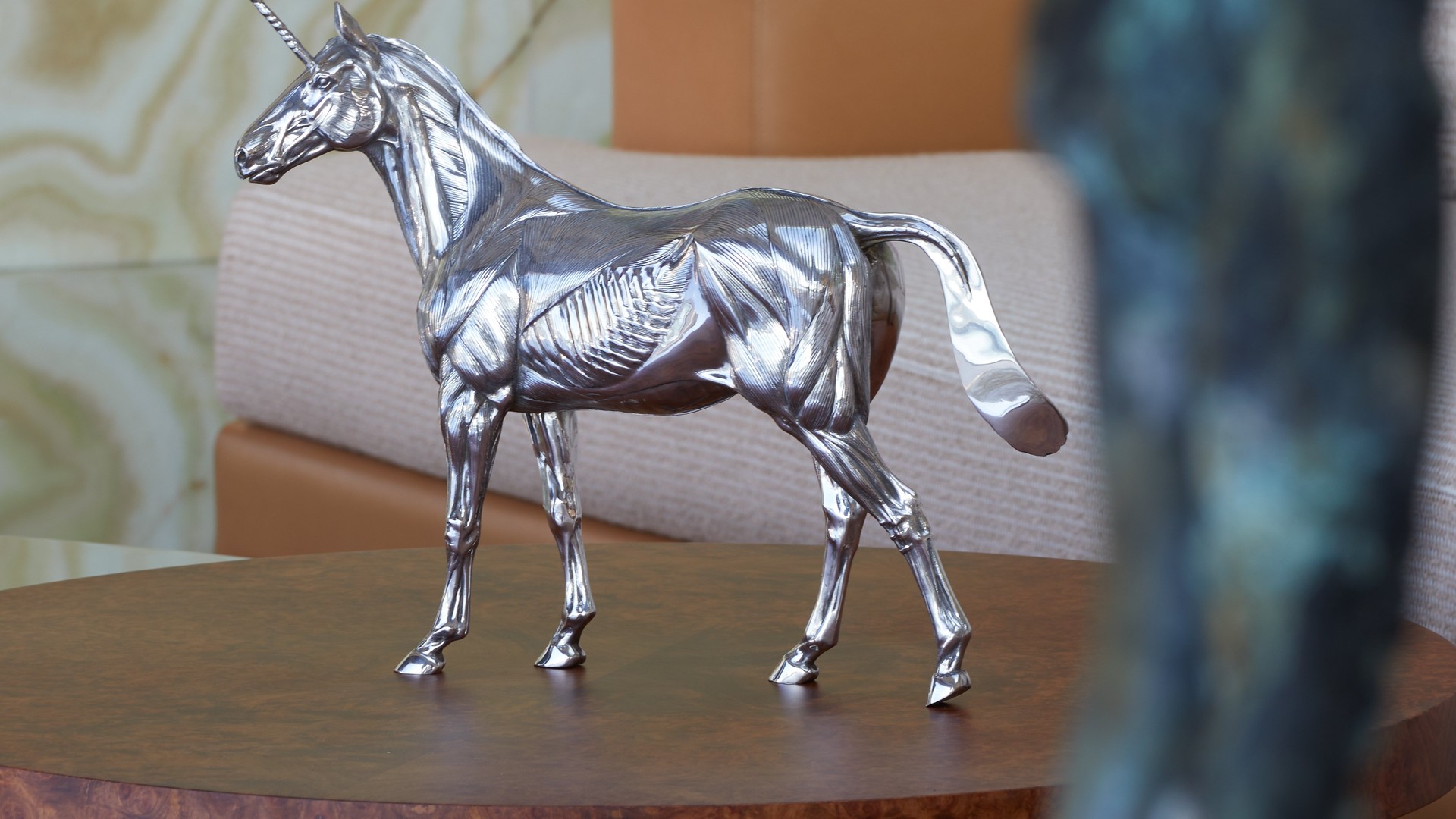 A sleek silver unicorn sculpture displayed on a rich wooden table against a backdrop of neutral-toned furniture and textured stone.