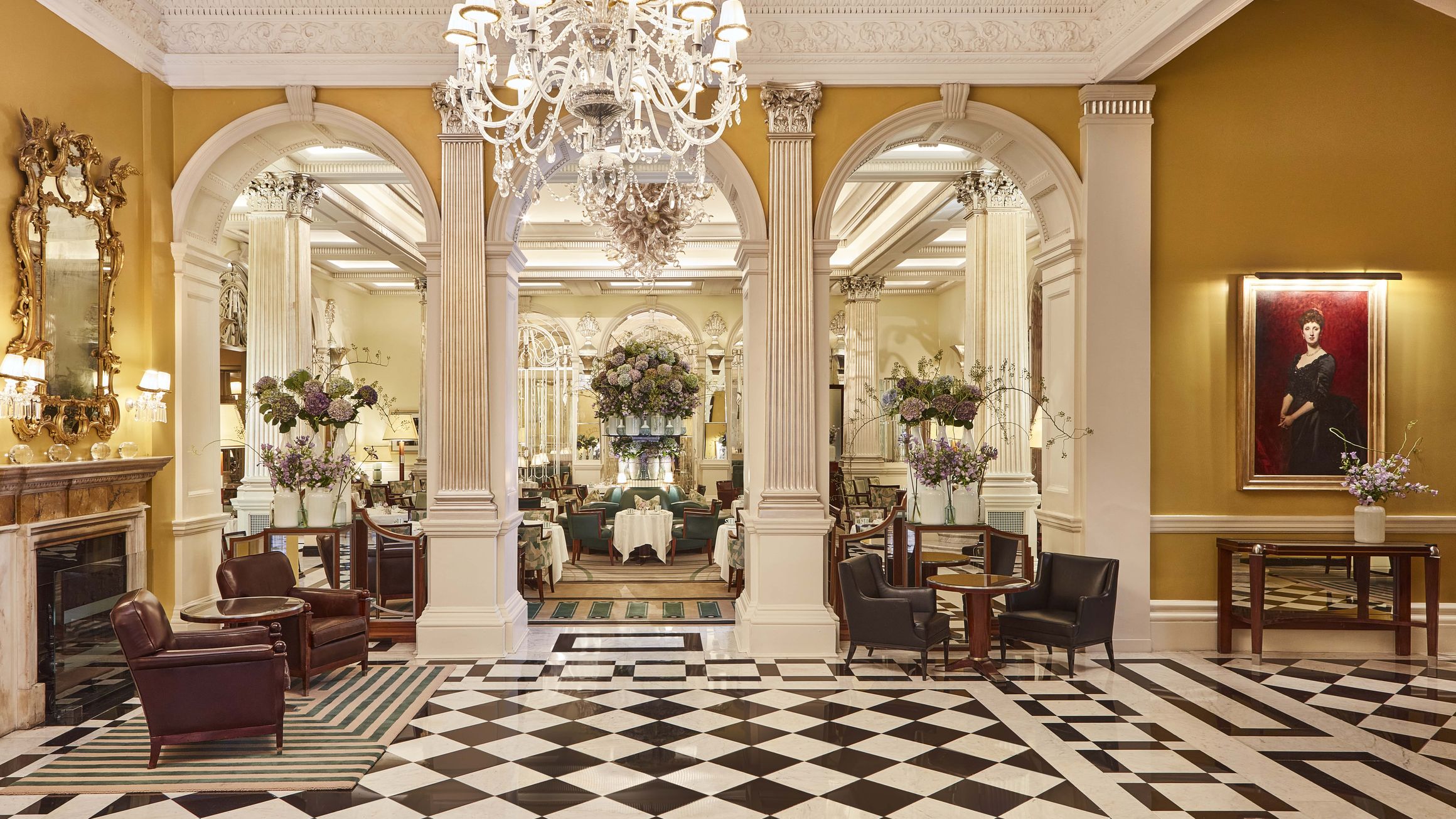 Hotel lobby with checkered marble flooring, chandeliers, classical columns, and floral arrangements, leading to a grand, mirrored tea lounge filled with guests.