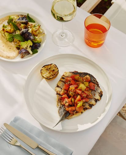 Grilled fish with tomato topping, charred lemon, roasted vegetables, and wine on a white tablecloth set for dining.