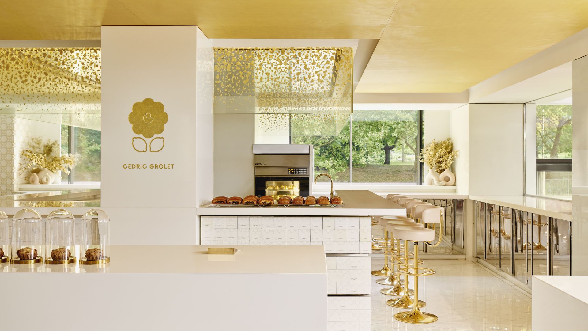 Minimalist pâtisserie interior in soft ivory and gold tones, featuring a central counter with glass-domed pastries, a row of golden bar stools, and floor-to-ceiling windows overlooking lush greenery.