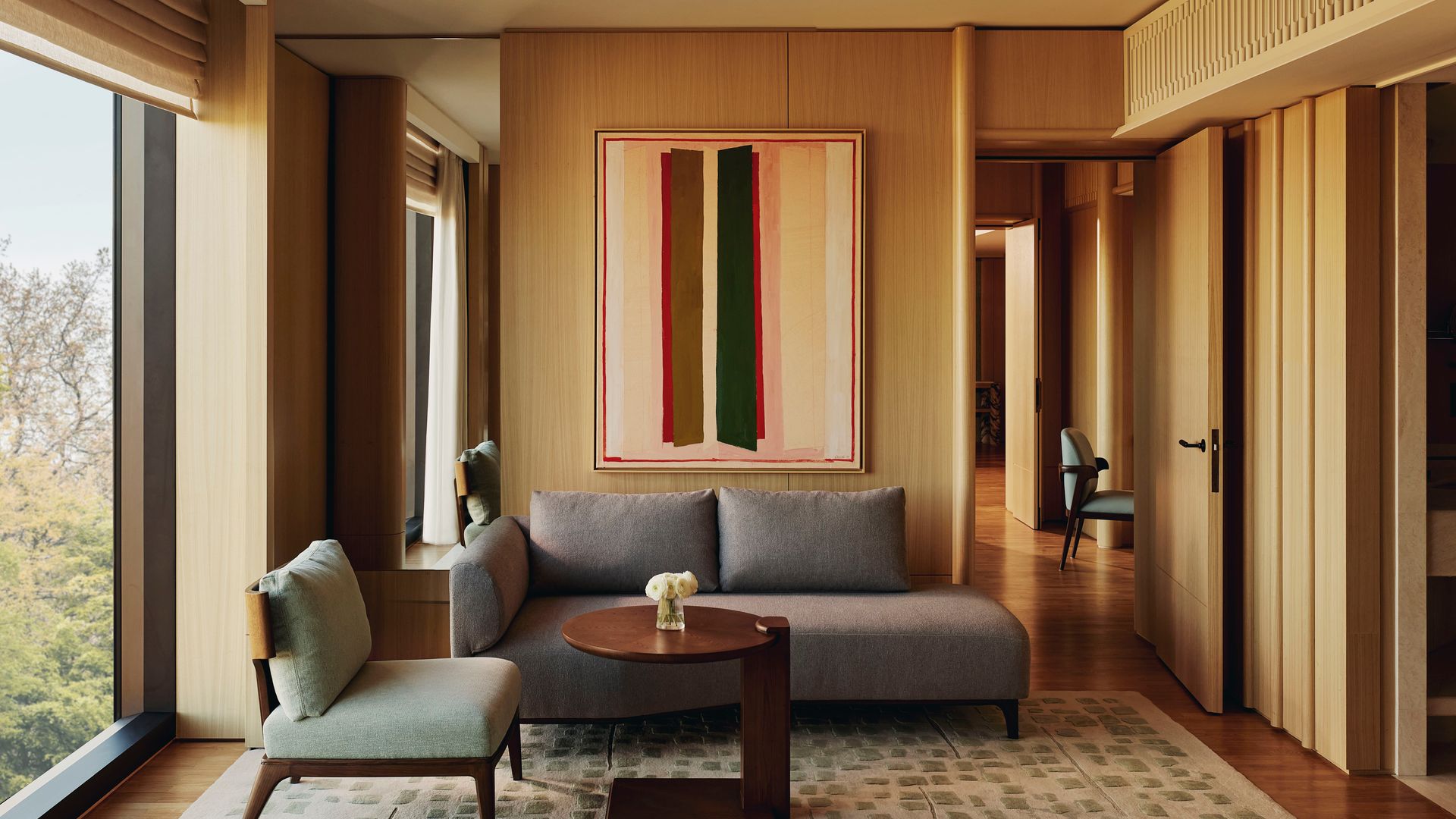 Modern suite living room with minimalist sofa, green accent chair, abstract wall art, and full-height windows framing treetop views.