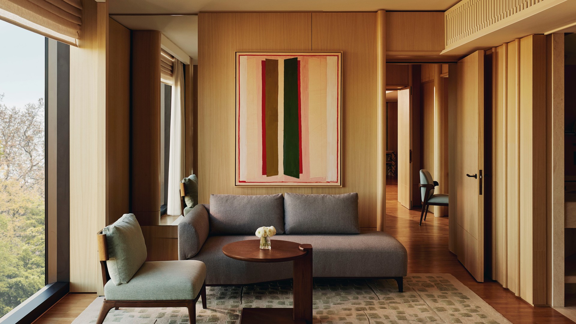 Modern suite living room with minimalist sofa, green accent chair, abstract wall art, and full-height windows framing treetop views.