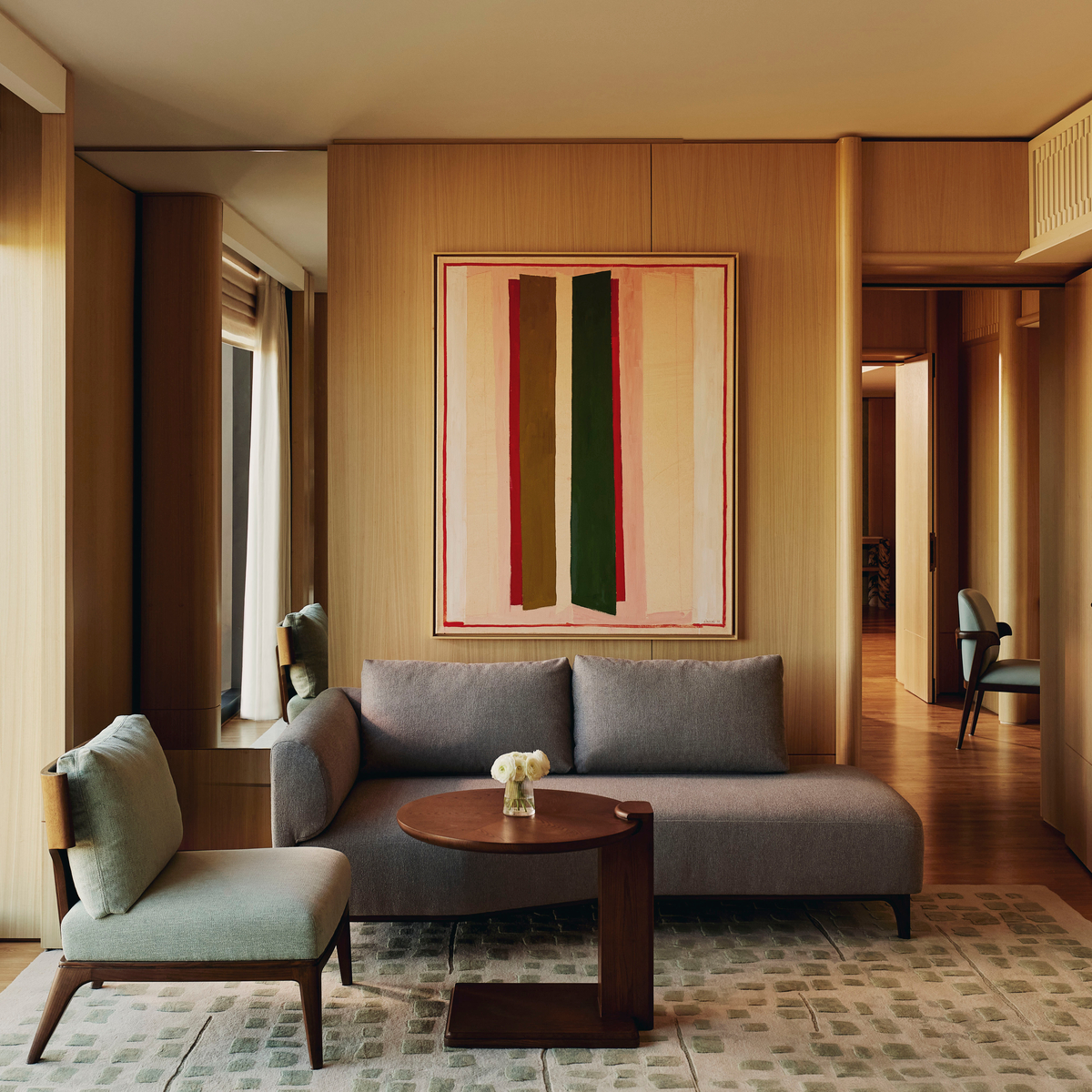 Modern suite living room with minimalist sofa, green accent chair, abstract wall art, and full-height windows framing treetop views.
