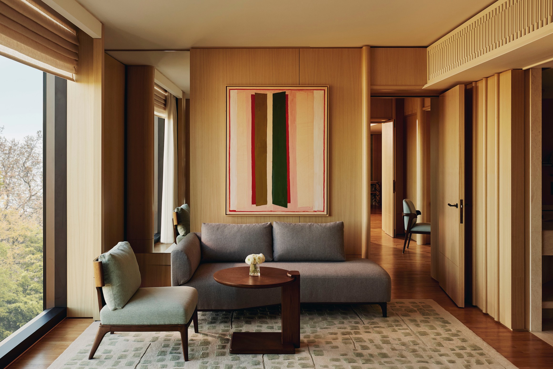 Modern suite living room with minimalist sofa, green accent chair, abstract wall art, and full-height windows framing treetop views.