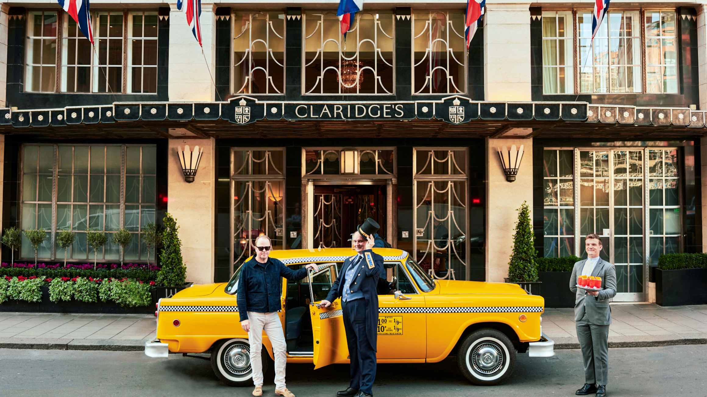 Doorman opens yellow taxi door for guest outside Claridge’s, with flags above entrance and staff member holding drinks