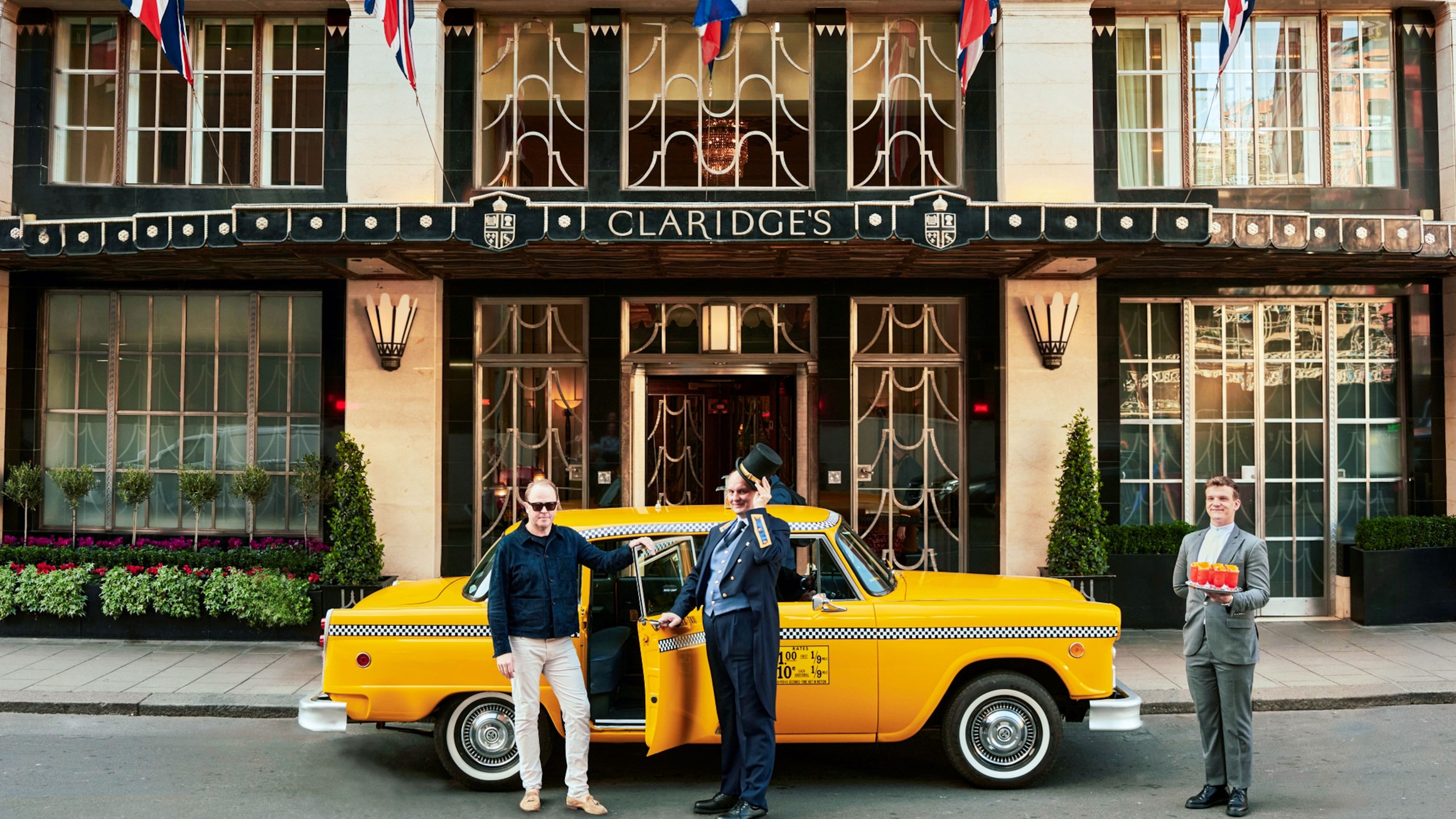 A man arrives at Claridge’s in a yellow taxi, greeted by a doorman and a staff member holding drinks.