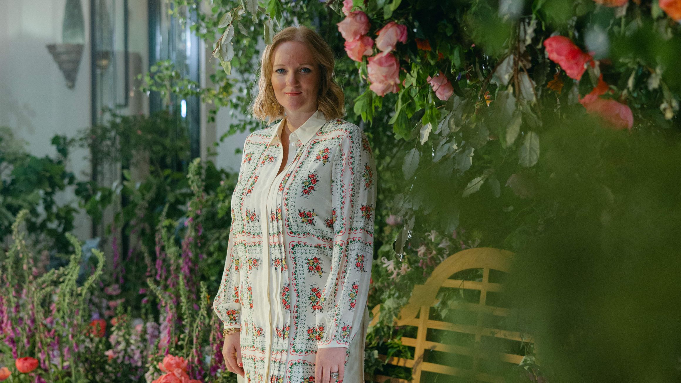 A woman stands gracefully in a floral-themed setting, surrounded by lush greenery and vibrant blossoms, exuding elegance in a patterned dress and espadrille wedges.