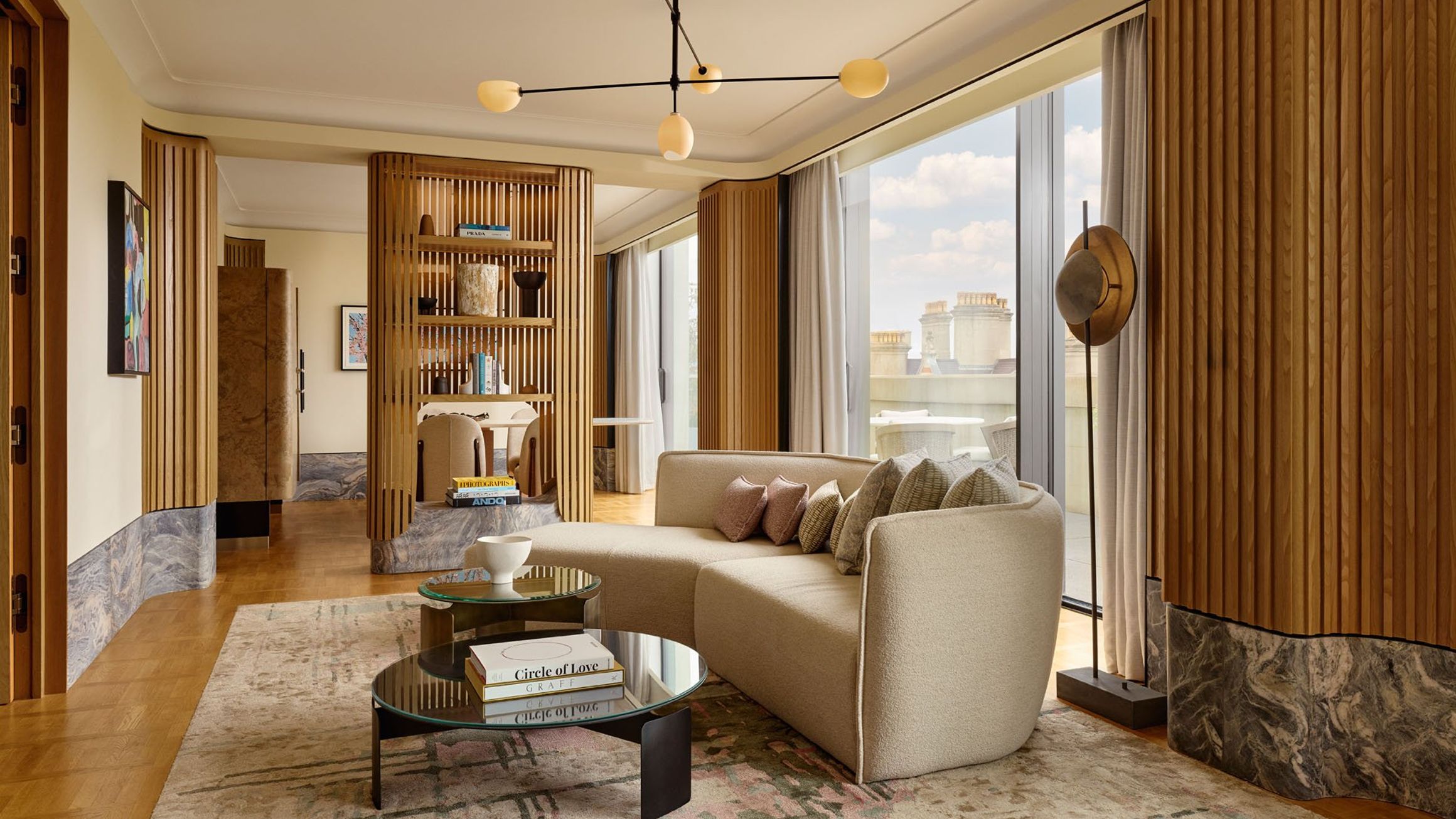 Light-filled penthouse living space with curved sofa, modern chandelier, sculptural shelving, and floor-to-ceiling windows opening onto a terrace with views over London’s rooftops.