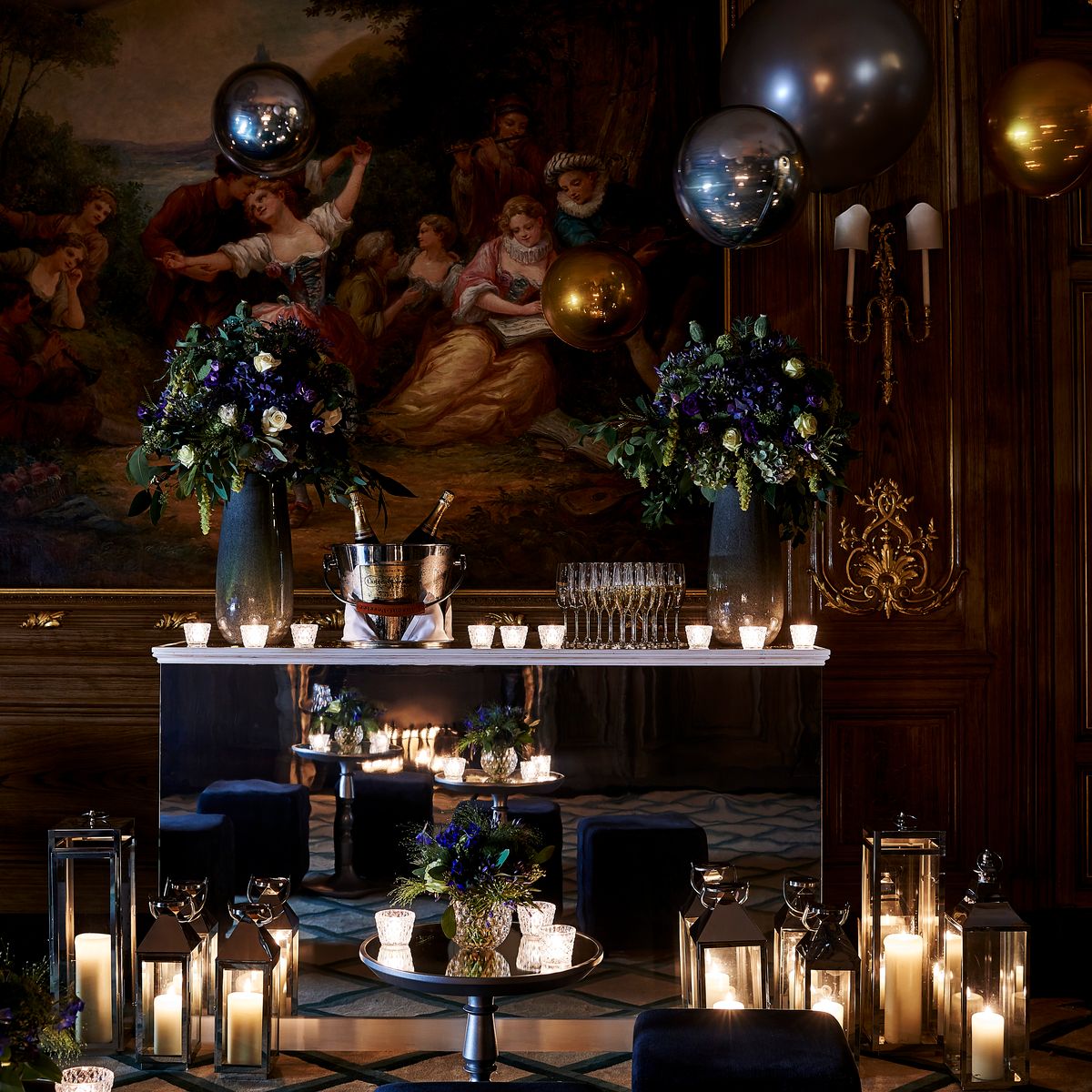 A glamorous party setup with floating balloons, luxurious florals, candlelit lanterns, and a mirrored bar in an ornate room.