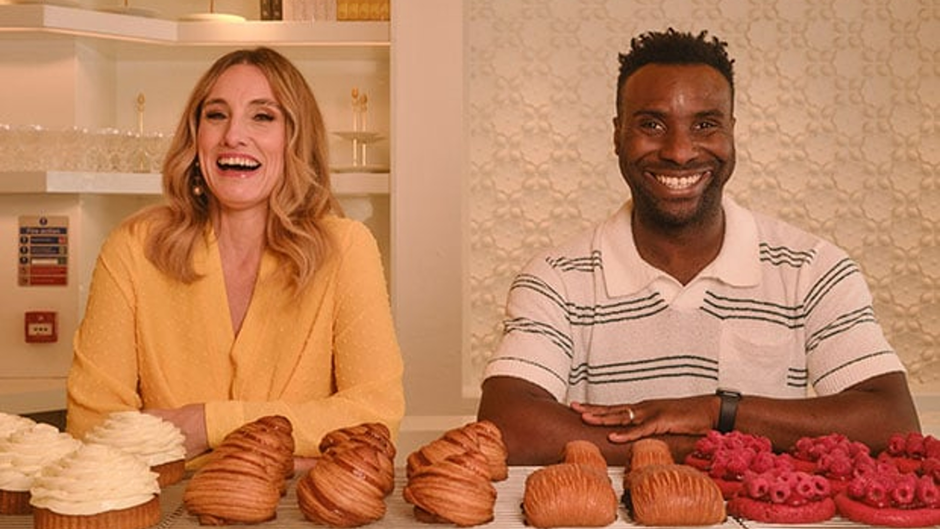 Two smiling guests seated behind a counter lined with artful pastries, including layered croissants and fruit tarts, set against a warm, minimalist interior with soft lighting.