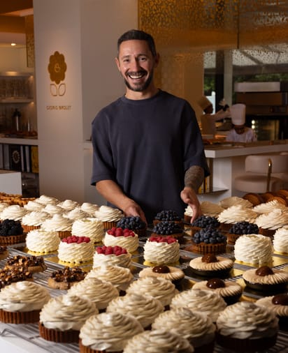 Pastry chef Cédric Grolet smiles behind a display of exquisite tarts, adorned with whipped cream swirls and fresh berries.