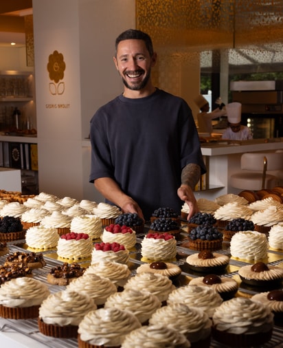 Pastry chef Cédric Grolet smiles behind a display of exquisite tarts, adorned with whipped cream swirls and fresh berries.