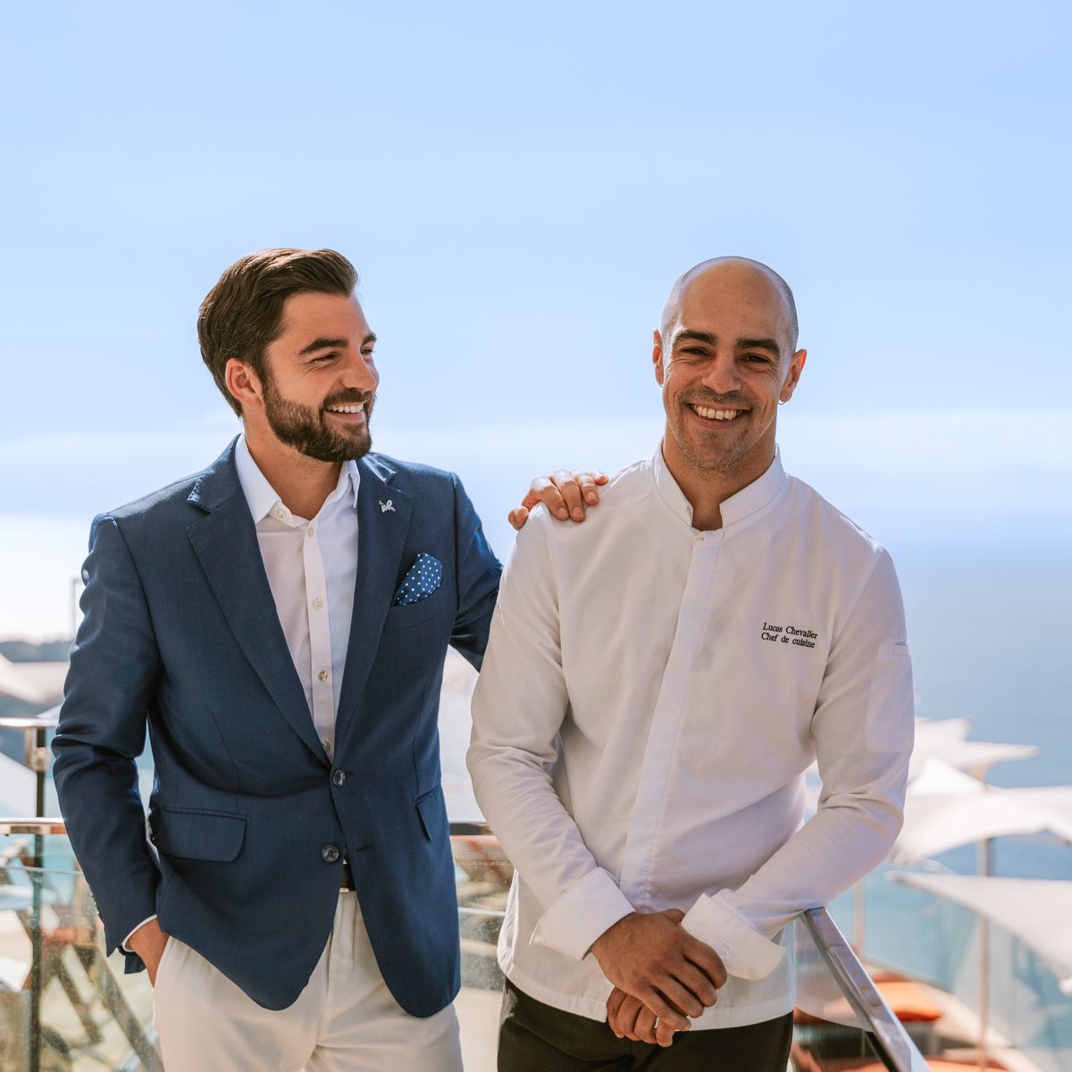 Two smiling professionals, a suited host and a chef in white, standing on a sunny terrace overlooking the sea.