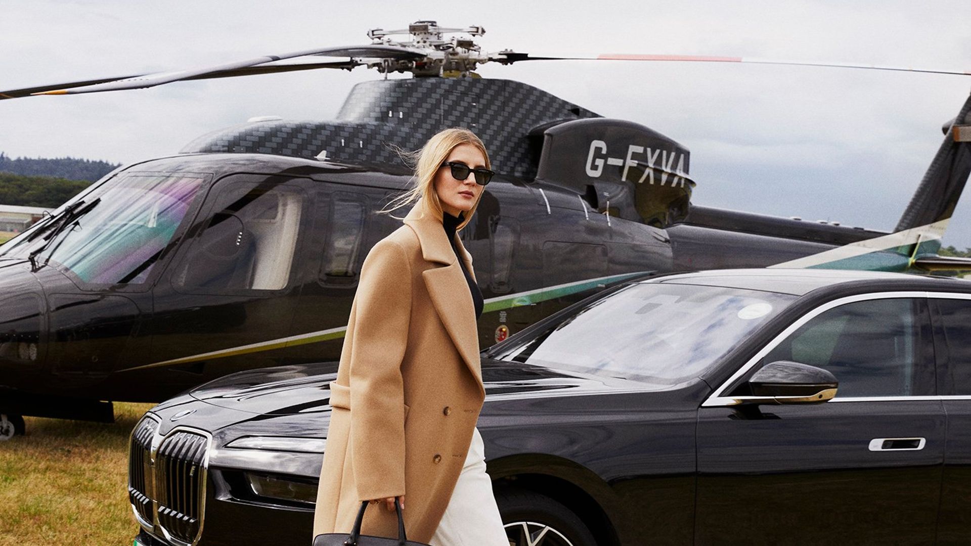 Woman in camel coat walking across grass between luxury car and black helicopter, suggesting high-end travel and exclusivity.