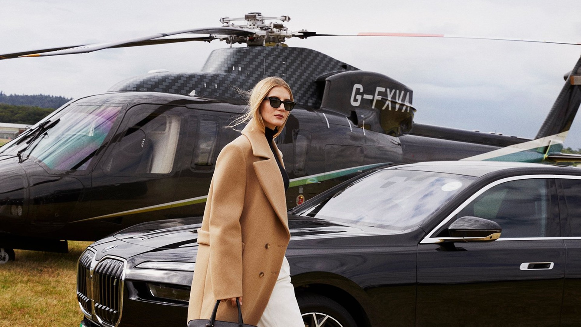 Woman in camel coat walking across grass between luxury car and black helicopter, suggesting high-end travel and exclusivity.
