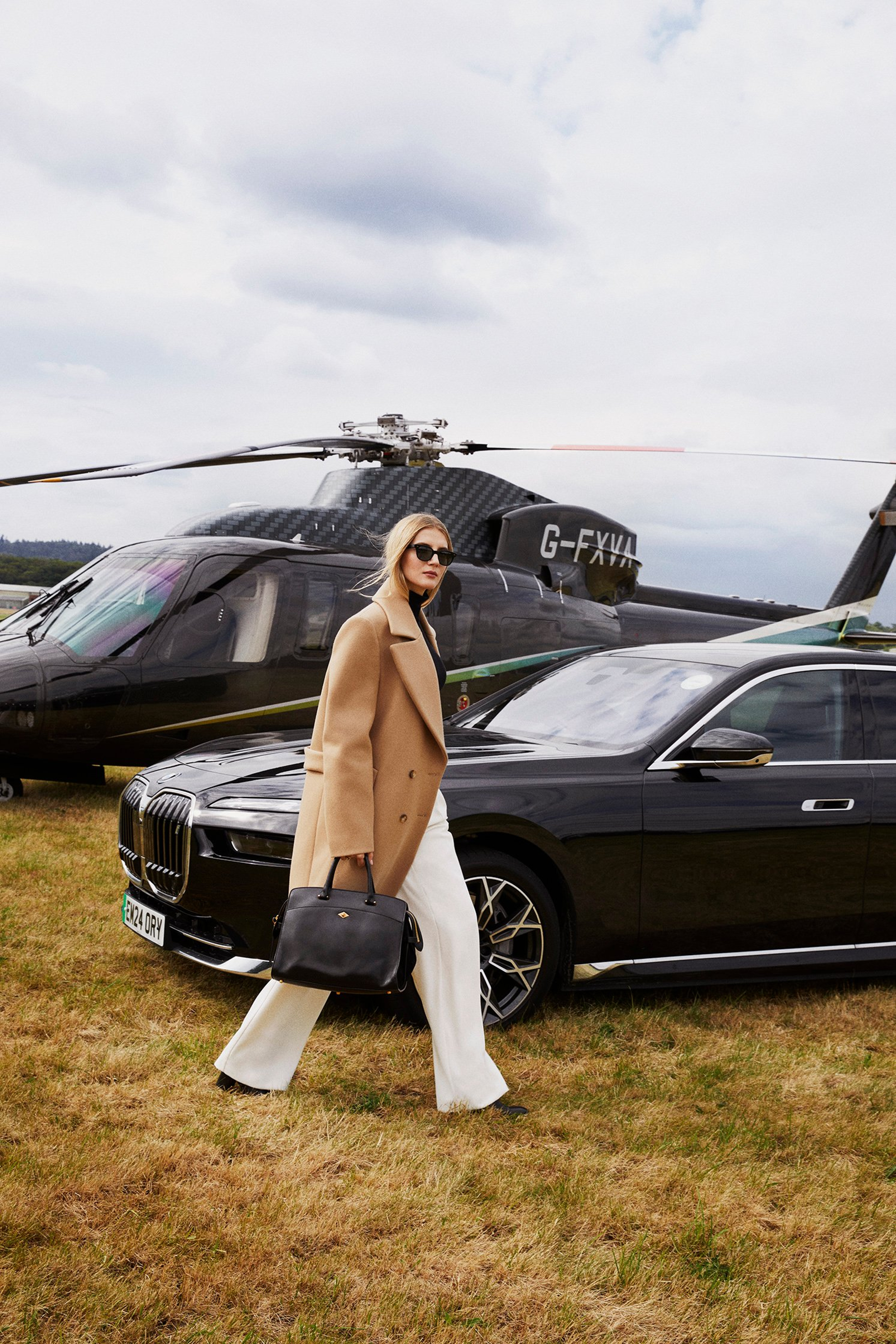 Woman in camel coat walking across grass between luxury car and black helicopter, suggesting high-end travel and exclusivity.